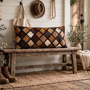 May include: A rectangular throw pillow featuring a geometric diamond pattern in alternating shades of dark brown, light brown, and cream. The pillow has a textured surface and is positioned horizontally on a wooden bench.