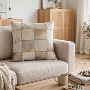 May include: A square throw pillow featuring a patchwork design of cream-colored hair-on hide and metallic gold foil-accented leather squares. The pillow is constructed with visible stitching connecting each individual square panel in a grid pattern.