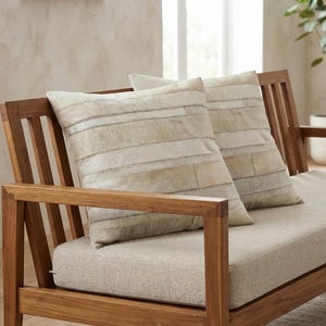 Peut inclure: Deux coussins carrés présentant un motif à rayures horizontales composé de panneaux en peau de vache beige clair et crème. Les coussins sont placés sur une structure de canapé en bois avec un coussin d'assise en tissu beige clair et un dossier à lattes.