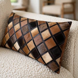 May include: A rectangular throw pillow featuring a patchwork design of cowhide squares in various shades of brown, tan, cream, and black. The squares are arranged in a diamond pattern and separated by thin, dark, straight lines across the surface.