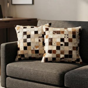 May include: Two square throw pillows featuring a patchwork design made of small, alternating squares of fur in shades of cream, tan, brown, and black. The pillows have a textured, multi-tonal appearance with a visible grain pattern across the surface.