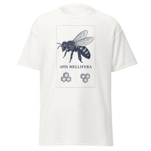 Puede incluir: Una camiseta blanca de manga corta con un gráfico rectangular central. El diseño incluye una ilustración detallada en tinta negra de una abeja, el texto APIS MELLIFERA y dos pequeños patrones hexagonales de panal encerrados en un borde punteado.
