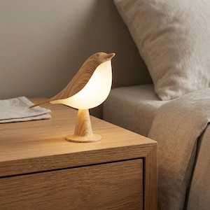 Puede incluir: Una lámpara de mesa con forma de pájaro que presenta un acabado de veta de madera clara en la cabeza, la cola y la base, con un cuerpo blanco mate iluminado que emite un brillo suave y cálido. La lámpara se apoya en una base circular.