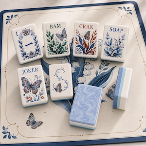 May include: A set of rectangular mahjong tiles featuring white surfaces with blue, green, and red botanical and butterfly illustrations. The tiles display text including 1, BAM, CRAK, SOAP, and JOKER, alongside a tile with a blue marbled pattern.