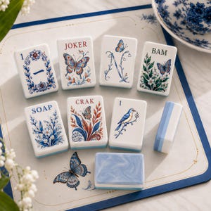 May include: A set of rectangular mahjong tiles featuring white surfaces with blue, orange, and green botanical, butterfly, and bird illustrations. The tiles include text labels such as JOKER, BAM, SOAP, and CRAK, alongside numerical markings and a blue marbled tile.