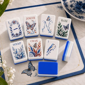 May include: A set of white rectangular Mahjong tiles featuring blue, red, and green botanical and butterfly illustrations. The tiles display text including JOKER, BAM, SOAP, and CRAK, alongside numerical markings and a blue bird on a branch.