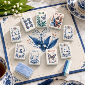 May include: A set of rectangular mahjong tiles featuring white surfaces with blue and red floral, butterfly, and numerical designs. The tiles include text such as BAM, CRAK, SOAP, and JOKER, alongside Chinese characters, arranged on a decorative mat.