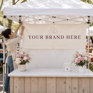 May include: A cream-colored rectangular banner with the text "YOUR BRAND HERE" in dark maroon lettering, featuring a pink line-art flourish above the text. The banner is mounted on a white pop-up canopy frame above a wooden market stall counter.