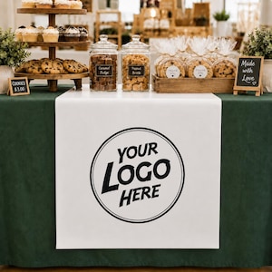 May include: A white table runner featuring a circular black logo placeholder with the text YOUR LOGO HERE, displayed on a dark green tablecloth. The table includes a tiered cupcake stand, two glass jars labeled Caramel Fudge and Peanut Brittle.