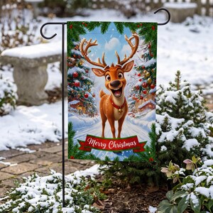 A festive Christmas-themed garden flag featuring a cartoon reindeer standing in a snowy landscape, with a "Merry Christmas" banner below.