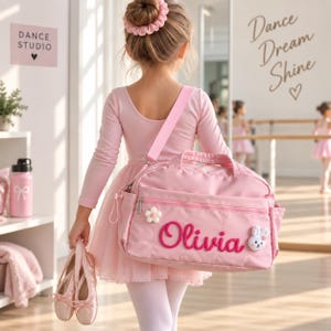 May include: A light pink duffel bag featuring the name Olivia in dark pink script, a white flower applique, and a small white rabbit face patch. The bag has a top handle, a shoulder strap, and a zippered front pocket, shown in a dance studio setting.