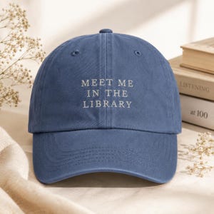 May include: A blue baseball cap featuring the embroidered white text "MEET ME IN THE LIBRARY" centered on the front panel. The cap has a curved brim, a button on the top, and stitched eyelets for ventilation, constructed from a solid fabric.