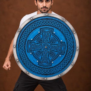 May include: A circular shield featuring a vibrant blue surface with intricate black Celtic knotwork patterns and a central cross design. The shield is framed by a metallic silver border accented with evenly spaced silver rivets along the outer edge.