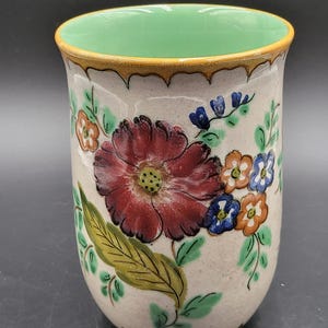 May include: A ceramic tumbler with a light beige base featuring a hand-painted floral design. The exterior displays a large dark red flower, smaller orange and blue blossoms, and green leaves. The interior is glazed in a solid light mint green.