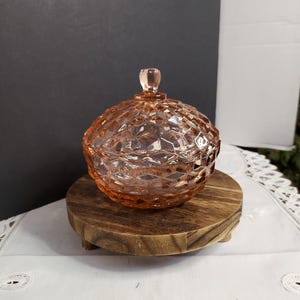 May include: A light pink glass candy dish with a matching lid, featuring a geometric diamond-cut pattern across the surface and a small, clear, faceted knob handle on top. The dish has a rounded, bowl-like shape and a translucent finish.