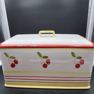 Peut inclure: Une boîte à pain rectangulaire en céramique avec une base blanche et un couvercle assorti doté d'une poignée jaune. L'avant est décoré de trois grappes de cerises rouges peintes avec des feuilles vertes, soulignées par des rayures horizontales.