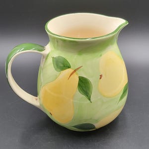 May include: A ceramic pitcher featuring a hand-painted design of yellow pears and green leaves against a light green wash background. The pitcher has a curved handle, a wide spout, and a cream-colored interior, showcasing a rounded body shape.