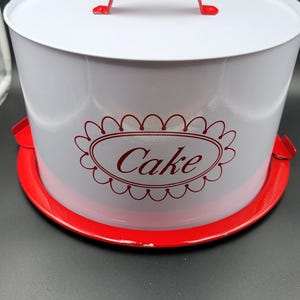 May include: A white metal cake carrier with a bright red circular base and a matching red arched handle on the lid. The side of the white cylindrical lid features a red decorative oval frame with the word Cake printed in a red script font.