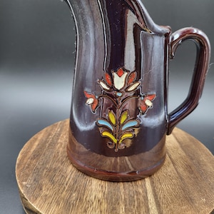 May include: A dark brown ceramic pitcher with a glossy finish and a curved handle. The front of the pitcher features a raised, hand-painted floral design in shades of white, red, yellow, and light blue, centered on the body of the vessel.