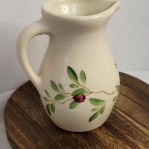 May include: A cream-coloured ceramic pitcher with a speckled finish and a single handle. The body of the pitcher is decorated with a hand-painted design featuring thin brown branches, green leaves, and a single dark red berry on the front.