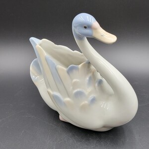 Peut inclure: Une figurine de cygne en porcelaine avec une finition blanche brillante et des accents bleu doux sur la tête et les plumes des ailes. Le cygne a un cou courbé, un bec rose pâle et un centre creux, servant de pot ou de plat décoratif.