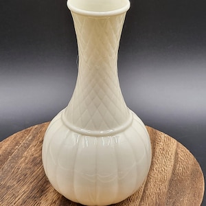 May include: A cream-colored ceramic vase with a glossy finish. The neck features a raised diamond lattice pattern, while the rounded base is decorated with vertical fluted ridges. The vase has a narrow opening and a distinct horizontal ring.