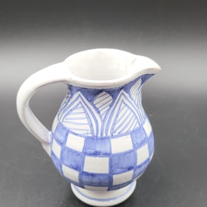 May include: A small ceramic pitcher featuring a hand-painted blue and white checkered pattern on the lower body and a series of blue triangular and striped motifs around the neck, complete with a curved handle and a flared pouring spout.