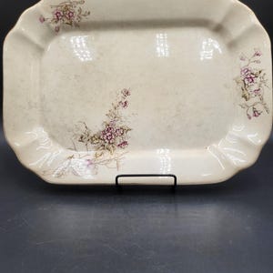 May include: A rectangular ceramic serving platter with a cream-colored base and scalloped edges. The platter features delicate floral illustrations in shades of pink, brown, and green, positioned in the upper left and right corners and the center.