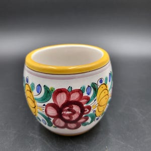 May include: A small, rounded ceramic cup with a white base, featuring hand-painted floral motifs in deep red, yellow, and green. The rim is accented with a solid yellow band, and the exterior displays symmetrical botanical patterns around the body.