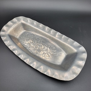 May include: A rectangular silver-toned metal tray with a ruffled, scalloped edge. The center of the tray features an embossed floral design with detailed petals and leaves, creating a textured surface across the base of the elongated dish.