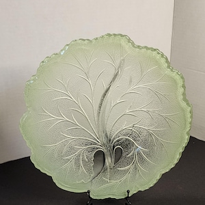 May include: A light green glass plate shaped like a large leaf with a textured surface and a prominent central vein. The edges of the plate are irregular and scalloped, mimicking the natural form of foliage. It rests on a black metal stand.