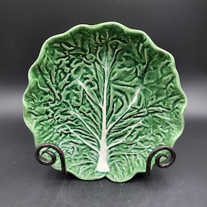 May include: A ceramic plate shaped like a green cabbage leaf with a prominent white central vein and textured, ruffled edges. The plate is displayed on a black metal stand with two curved supports that hold the dish in an upright position.