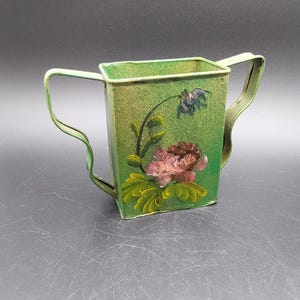 May include: A rectangular green metal container with two curved handles on the sides. The front surface features a hand-painted design of a large pink flower with green leaves and a smaller blue flower bud extending from a thin stem on a textured finish.