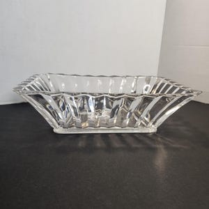 May include: A clear, rectangular glass bowl featuring a faceted, geometric design with vertical ridges along the sides and a wide, angled rim. The bowl has a smooth, flat base and a transparent, reflective surface that catches light.