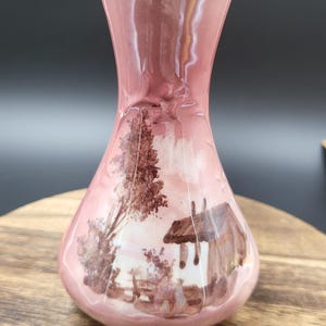 May include: A pink ceramic vase with a flared neck and a gold-toned rim. The body of the vase features a brown painted landscape scene depicting a small cottage, a tall tree, and foliage. The surface has a glossy finish with a smooth, curved shape.