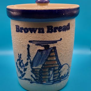Puede incluir: Un tarro cilíndrico de cerámica con tapa, que presenta un acabado moteado en color blanco roto y un borde azul oscuro. El texto "Brown Bread" está impreso en azul, junto a una ilustración de líneas azules de una pequeña cabaña.