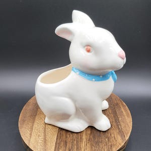 Może przedstawiać: Biała ceramiczna doniczka w kształcie królika z błyszczącym wykończeniem. Królik ma różowe oczy i nos oraz jasnoniebieską obrożę w małe białe kropki wokół szyi. Pusty otwór na rośliny znajduje się na grzbiecie figurki.