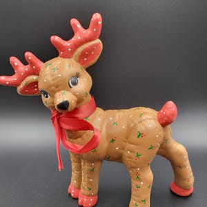 Könnte beinhalten: Eine hellbraune Rentierfigur mit roten Geweihen, roten Hufen und einem roten Schwanz. Der Körper ist mit kleinen grünen Blattmustern, roten Beeren und weißen Punkten verziert. Ein rotes Satinband ist um den Hals des Rentiers gebunden.