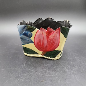 May include: A small, cream-colored metal planter with a scalloped top edge and a black interior. The exterior features hand-painted floral designs, including a prominent red tulip, green leaves, and blue petals against a light cream background.