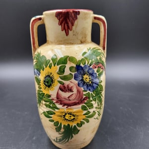 Puede incluir: Un jarrón de cerámica de color crema con dos asas, decorado con motivos florales pintados a mano en amarillo, azul y rosa, rodeados de hojas verdes. El borde y las asas están acentuados con pintura marrón rojiza y tiene un acabado brillante.