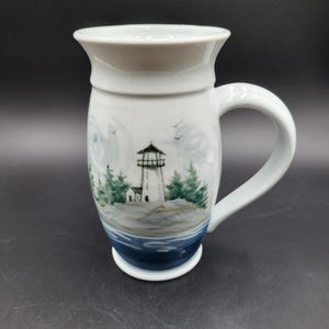 Puede incluir: Una taza de cerámica blanca con borde acampanado y asa curva. El exterior presenta una escena pintada a mano de un faro en una costa rocosa rodeado de pinos verdes, con agua azul en la base y pequeños pájaros en el cielo.