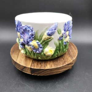 Op de afbeelding: Een witte keramische kom met een verhoogd, driedimensionaal reliëfontwerp van blauwe en paarse irissen met groene bladeren en gele accenten. De kom heeft een gladde, glanzende afwerking en is rondom versierd met gedetailleerde bloemmotieven.