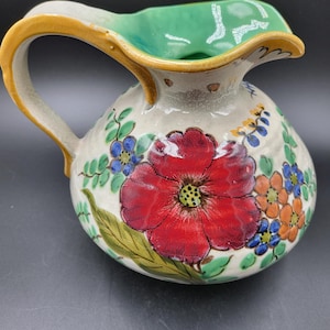 May include: A ceramic pitcher with a cream-colored base, featuring a large central red flower, smaller blue and orange blossoms, and green leaves. The handle and rim are glazed in a mustard yellow, while the interior is a bright mint green.