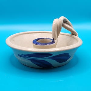 Puede incluir: Un candelabro de cerámica color crema con acabado moteado, que presenta una copa central para vela con borde azul, un asa trenzada y un diseño de hojas azules pintado a mano alrededor del exterior de la base poco profunda.
