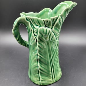 Puede incluir: Una jarra de cerámica verde brillante con un diseño de hoja moldeada que imita la textura y las venas de una hoja de col grande. El recipiente tiene un asa curva en un lado y un pico vertedor ancho y festoneado en el borde superior.