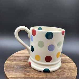 May include: A cream-colored ceramic mug featuring a repeating pattern of multicolored polka dots in shades of red, blue, green, and yellow. The mug has a curved handle and a raised base, with a smooth, glossy finish throughout the exterior surface.