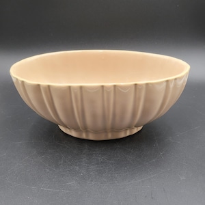 May include: A light beige ceramic bowl featuring a rounded shape and a series of vertical fluted indentations along the exterior surface. The bowl has a smooth, glossy finish and sits on a slightly raised circular base, presenting a uniform color.
