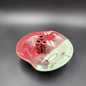 Könnte beinhalten: Eine Blumenigel-Schale aus Keramik mit glänzender Oberfläche und einem zweifarbigen Design in Dunkelrot und Hellgrün. In der Mitte der Schale befinden sich drei aufrechte zylindrische Röhren zum Halten von Stielen, verziert mit einem gemalten Blumenmotiv.