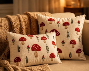 Op de afbeelding: Twee gebroken witte sierkussens met een geborduurd patroon van rode paddenstoelen met beige stelen, afgewisseld met kleine groene varenachtige takjes en kleine rode besjes. De rechthoekige en vierkante kussens hebben een textuurafwerking.
