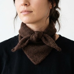 Op de afbeelding: Een donkerbruine gebreide halsdoek met een driehoekige vorm en een textuur van gebreid garen. Het accessoire is aan de voorzijde in een eenvoudige knoop gestrikt, rust rond de nek en wordt gedragen over een effen zwart T-shirt.